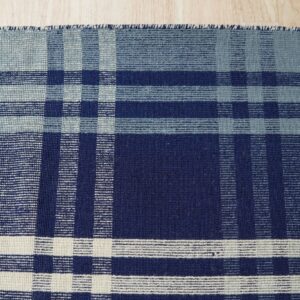A flatwoven plaid rug in shades of navy, slate blue, and cream rests on light wood flooring.