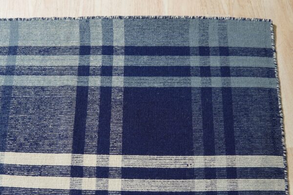 A flatwoven plaid rug in shades of navy, slate blue, and cream rests on light wood flooring.