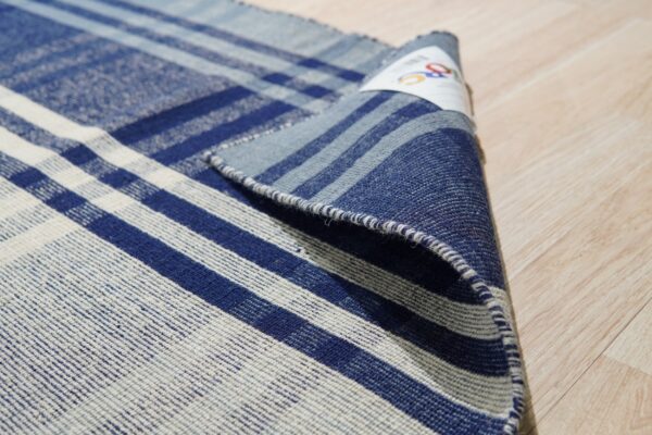 Partially rolled blue and off-white flatweave rug with a plaid pattern resting on a light wood floor.