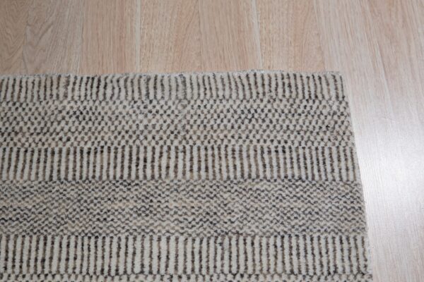 The edge of a beige rug with thick textured stripes and geometric patterns rests on light wood planks.