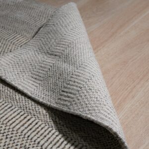 A low-pile beige rug with a dark chevron pattern is folded on a light wood floor.