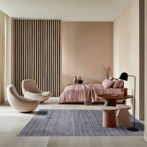 A textured gray area rug covers light wood floors in a modern bedroom with pink bedding and cream chairs.