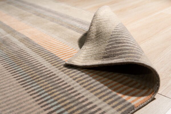 A low-pile rug with geometric stripe patterns in brown, tan, orange, and light blue is curled on light wood flooring.