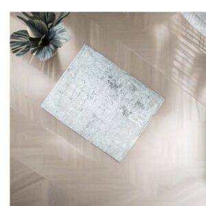 A light grey abstract rug rests diagonally on blonde herringbone wood flooring near a dark green plant.