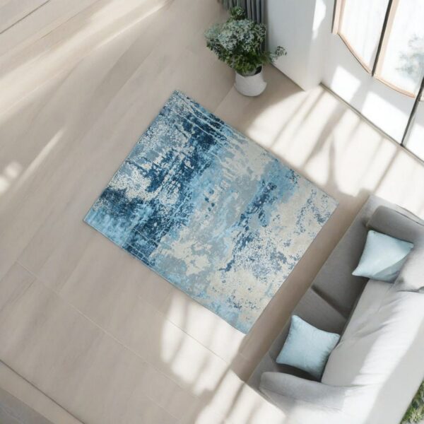 Abstract blue and gray distressed rug placed on light flooring next to a gray sofa and potted plant.