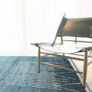 Blue rug with white speckles and horizontal stripes under a wooden chair by sheer curtains.