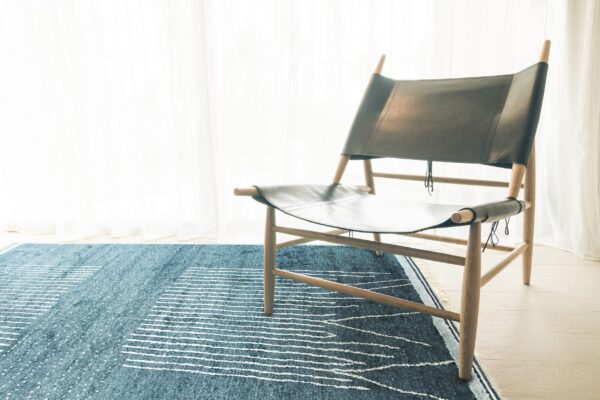 Blue rug with white speckles and horizontal stripes under a wooden chair by sheer curtains.
