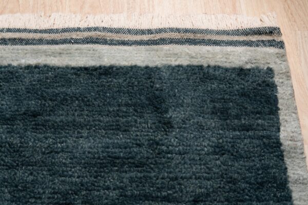 Dark teal shag rug edge with light gray border, striped flatweave bands, and cream fringe on a wood floor.