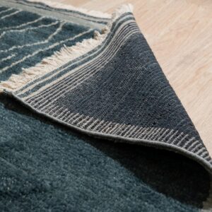 Teal high-pile rug with white stripes folded over, revealing striped woven backing and fringe on wood floors.