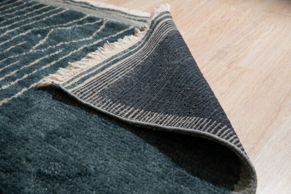 Teal high-pile rug with white stripes folded over, revealing striped woven backing and fringe on wood floors.