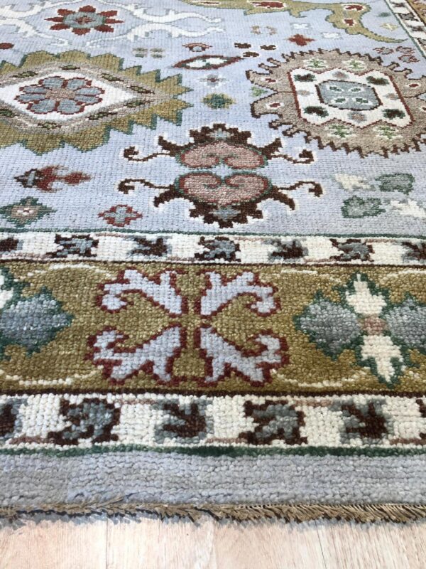 Light blue traditional rug with olive and rust geometric patterns resting on light wood flooring.