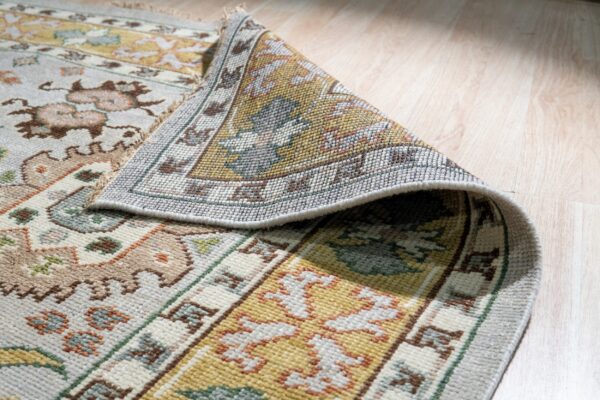 A dense, multicolored floral and geometric rug is partially rolled on blonde wood flooring.