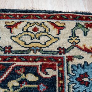 Corner detail of a patterned rug showing cream, navy, red, and gold colors on light wood flooring.