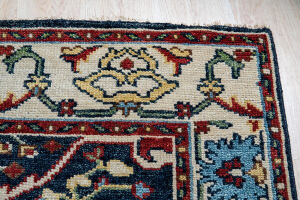Corner detail of a patterned rug showing cream, navy, red, and gold colors on light wood flooring.
