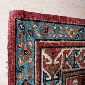 Textured red and teal rug with geometric borders rests on blonde wood plank flooring.
