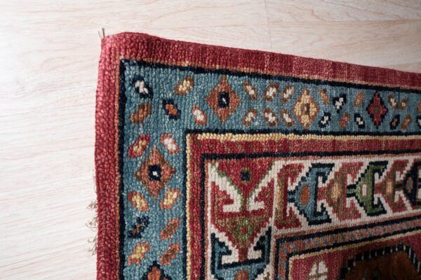 Textured red and teal rug with geometric borders rests on blonde wood plank flooring.