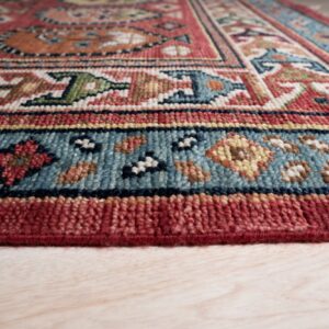 Geometric rug pattern in red, teal, tan, and blue, viewed at the bound edge on light wood planks.