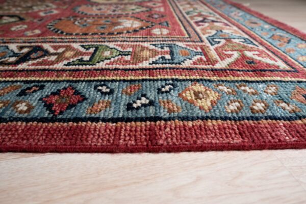 Geometric rug pattern in red, teal, tan, and blue, viewed at the bound edge on light wood planks.