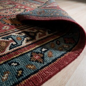 The textured, multicolored edge of a traditional rug is curled upward, revealing thick pile on light wood.