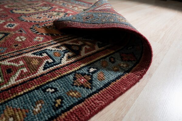 The textured, multicolored edge of a traditional rug is curled upward, revealing thick pile on light wood.