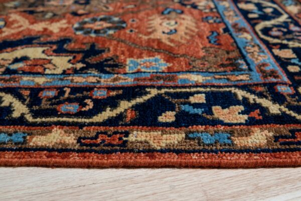 Close-up of a medium pile rug border in navy, rust, and beige, resting on light wood grain flooring.