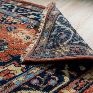 Navy, rust, and beige patterned rug with fringe, partially rolled back on light wood planks.