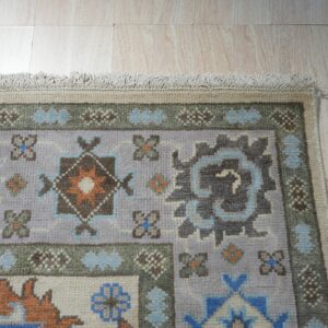 Lavender and blue patterned rug with geometric motifs and fringe rests on light wood plank flooring.