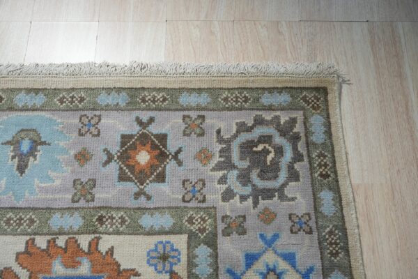 Lavender and blue patterned rug with geometric motifs and fringe rests on light wood plank flooring.