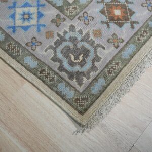 Geometric rug corner featuring lavender, blue, and sage green patterns with fringe on pale wood planks.