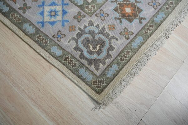 Geometric rug corner featuring lavender, blue, and sage green patterns with fringe on pale wood planks.
