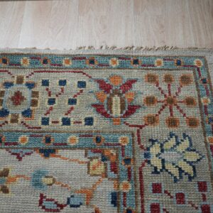 Traditional beige rug with red, teal, and orange geometric patterns rests on light wood grain floor.