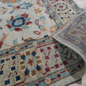 Grey rug featuring traditional blue, red, and orange patterns is shown partially rolled up on light wood floor.
