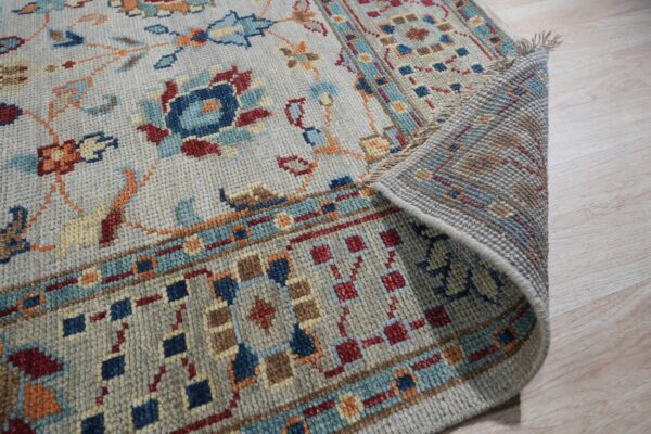 Grey rug featuring traditional blue, red, and orange patterns is shown partially rolled up on light wood floor.