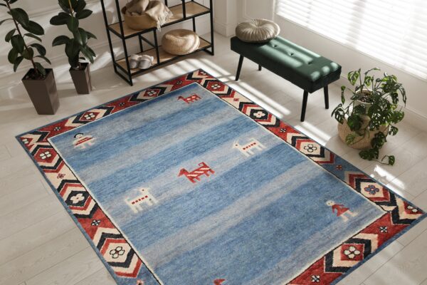 Light blue rug featuring abrash stripes, primitive figures, and a red geometric border in a sunlit room.