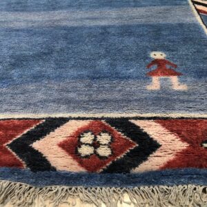 Textured blue rug with stylized figures and a red and black geometric border rests on wood flooring.