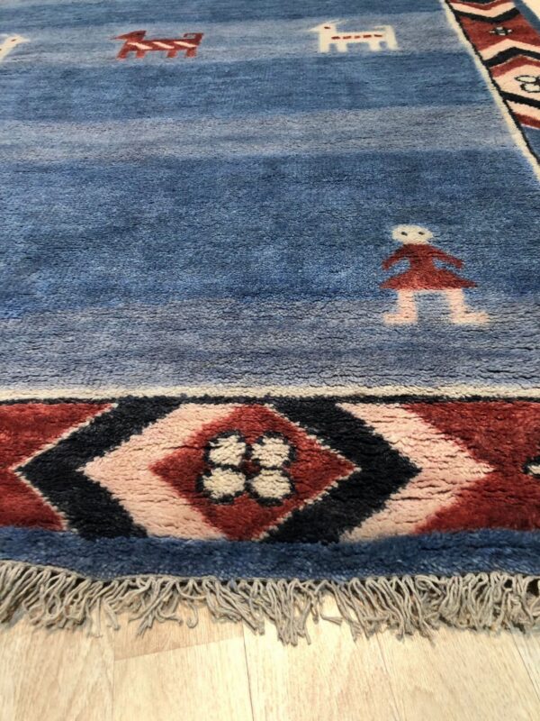 Textured blue rug with stylized figures and a red and black geometric border rests on wood flooring.