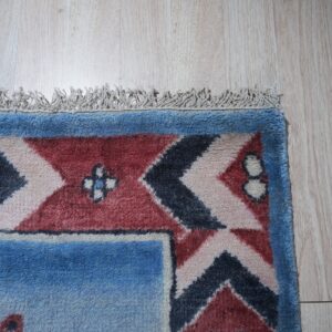 High-pile rug corner displaying blue, red, and navy geometric patterns and fringe on light wood flooring.
