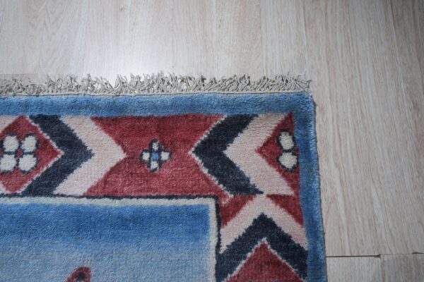 High-pile rug corner displaying blue, red, and navy geometric patterns and fringe on light wood flooring.