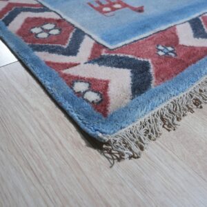 Textured blue and dusty red patterned rug corner with fringe lies on light wood laminate flooring.
