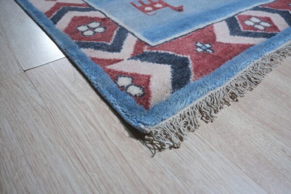 Textured blue and dusty red patterned rug corner with fringe lies on light wood laminate flooring.