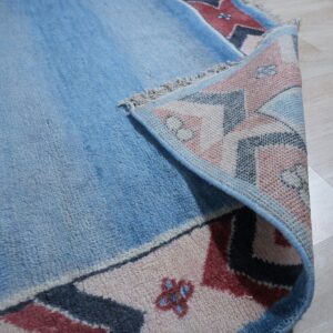 Blue high-pile rug with geometric pink, red, and navy border folded on light wood flooring.