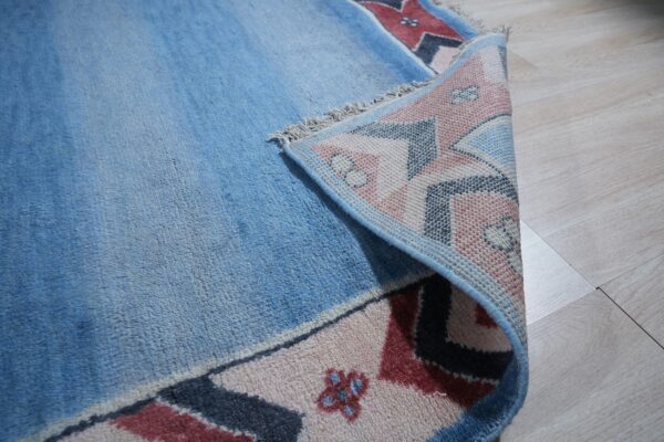 Blue high-pile rug with geometric pink, red, and navy border folded on light wood flooring.