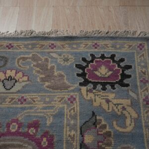 Corner of a blue and gray patterned rug featuring tassels and magenta accents on light wood flooring.