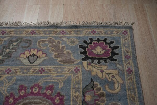 Corner of a blue and gray patterned rug featuring tassels and magenta accents on light wood flooring.