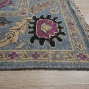 Blue-gray patterned rug featuring magenta, black, and yellow motifs, with fringe visible on light wood flooring.