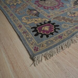 A traditional blue rug with yellow and magenta floral motifs rests on light blonde plank flooring.