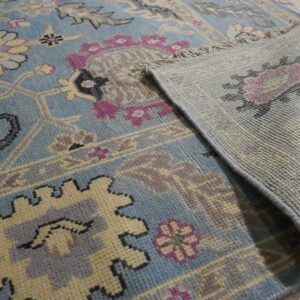 A blue, cream, and magenta traditional rug is folded over, showing the backing on light wood flooring.