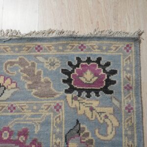 Blue patterned rug with fringed edge displays purple, black, and gray motifs on light wood planks.