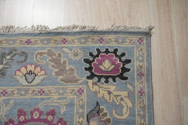Blue patterned rug with fringed edge displays purple, black, and gray motifs on light wood planks.