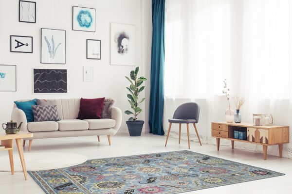 A light blue rug with a traditional medallion pattern anchors a brightly lit living room with a beige sofa and wooden furniture.